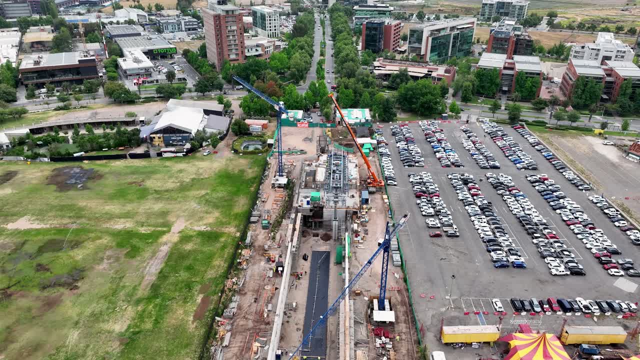 Forward drone aerial of urban construction site with cranes, machinery, large parking lot with cars, and commercial buildings backdrop
