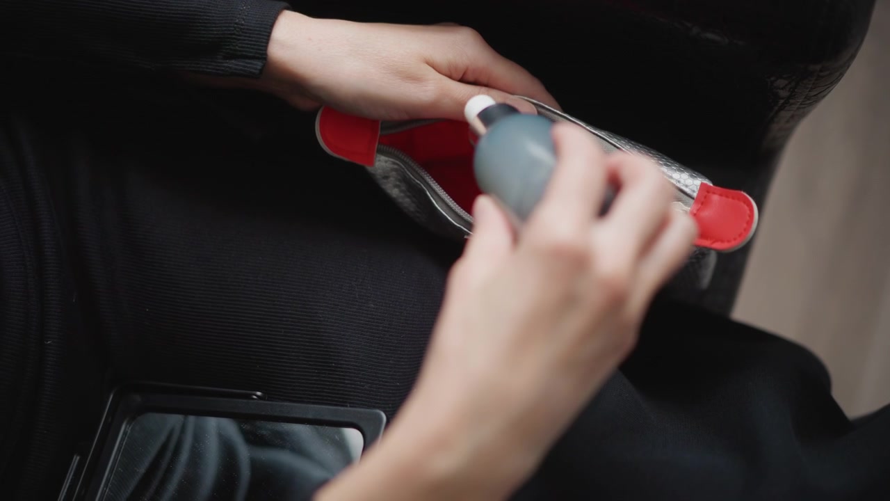 Primer plano de las manos de una mujer abriendo y cerrando la cremallera de un bolso de mano plateado texturizado con detalles rojos, revelando su interior. La escena se centra en el movimiento de la mano alrededor del bolso en un ambiente interior.