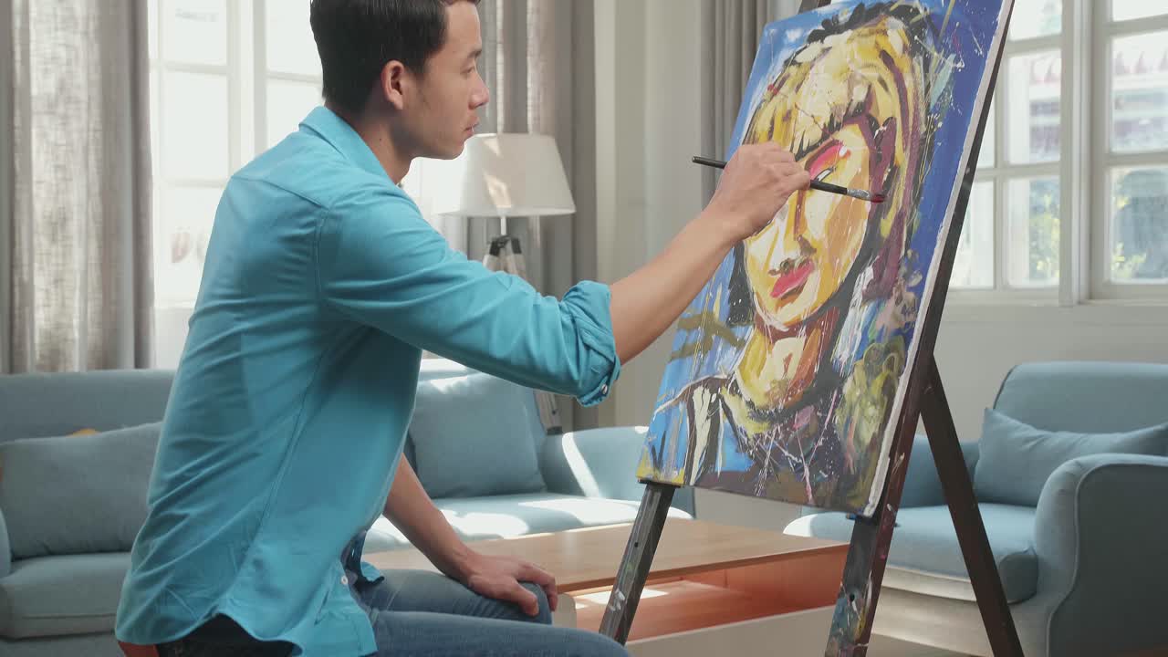 Medium Shot Side View Of An Asian Artist Man Sitting On Round Wooden Chair Without Backrest Holding Paintbrush And Mixing It With Colour Before Painting A Girl'S Hair On The Canvas