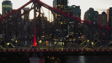 Futuristic-view-aerial-tracking-shot-along-famous-story-bridge-with-traffic-crossing-the-river-and-illuminated-cyberpunk-style-downtown-cityscape-background-at-sunset-golden-hour,-Brisbane-city