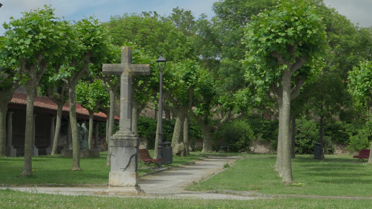 Toma estática de un monumento de cruz de piedra en un jardín de iglesia arbolado en Luanco, España. Césped verde, farola y arcada de iglesia histórica visibles