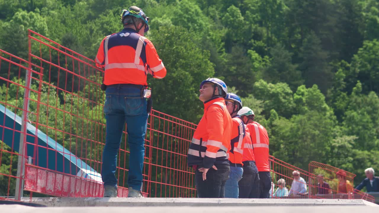 Workers in safety gear coordinate bridge rigging at a sunny construction site in rural France