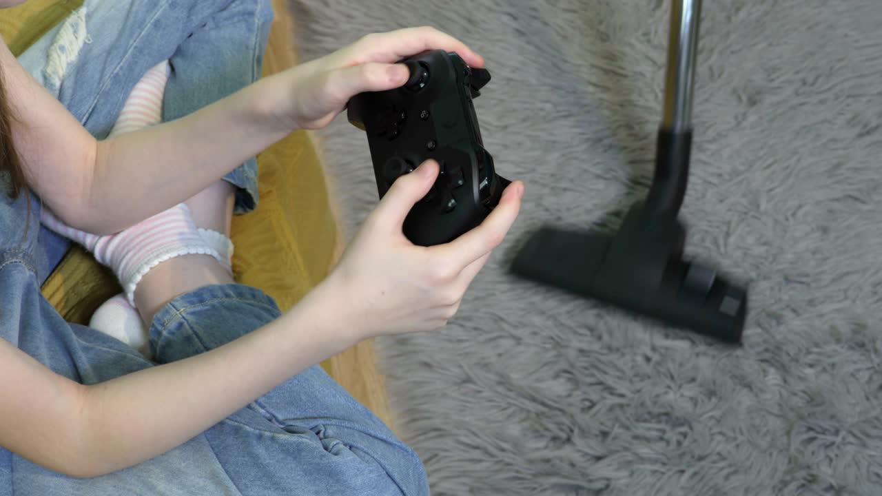 딸은 조이스틱을 들고 비디오를 연주하고 어머니는 뒤에서 진공청소기를 사용합니다.