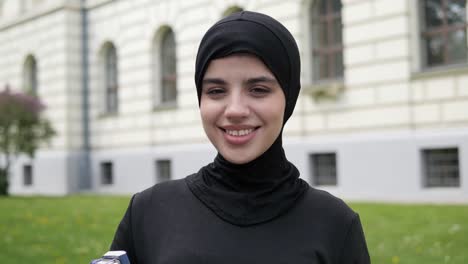 portrait of beautiful young muslim girl student in black traditional hijab looking straight to camera and smiling. close up of female pretty face with charming smile standing outside university.