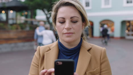 retrato en cámara lenta de una elegante mujer de negocios rubia en traje usando una aplicación de redes sociales de teléfono inteligente enviando mensajes de texto en un fondo urbano