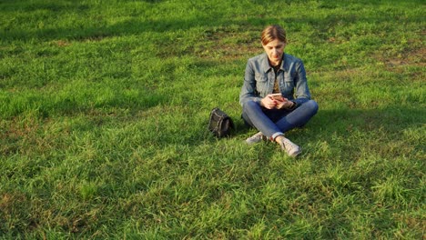 young woman spends time relaxing in the park. she sits on the green grass and reads from her phone screen.