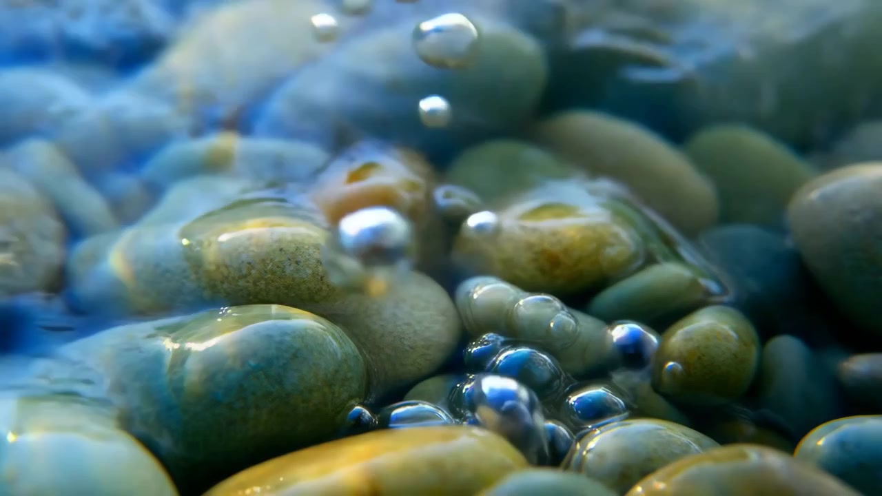 Un movimento ravvicinato mostra bolle sferiche che si staccano da ciottoli di fiume lisci e salgono attraverso acqua limpida e poco profonda, con un leggero luccichio, delicati riflessi e un'atmosfera naturale serena.
