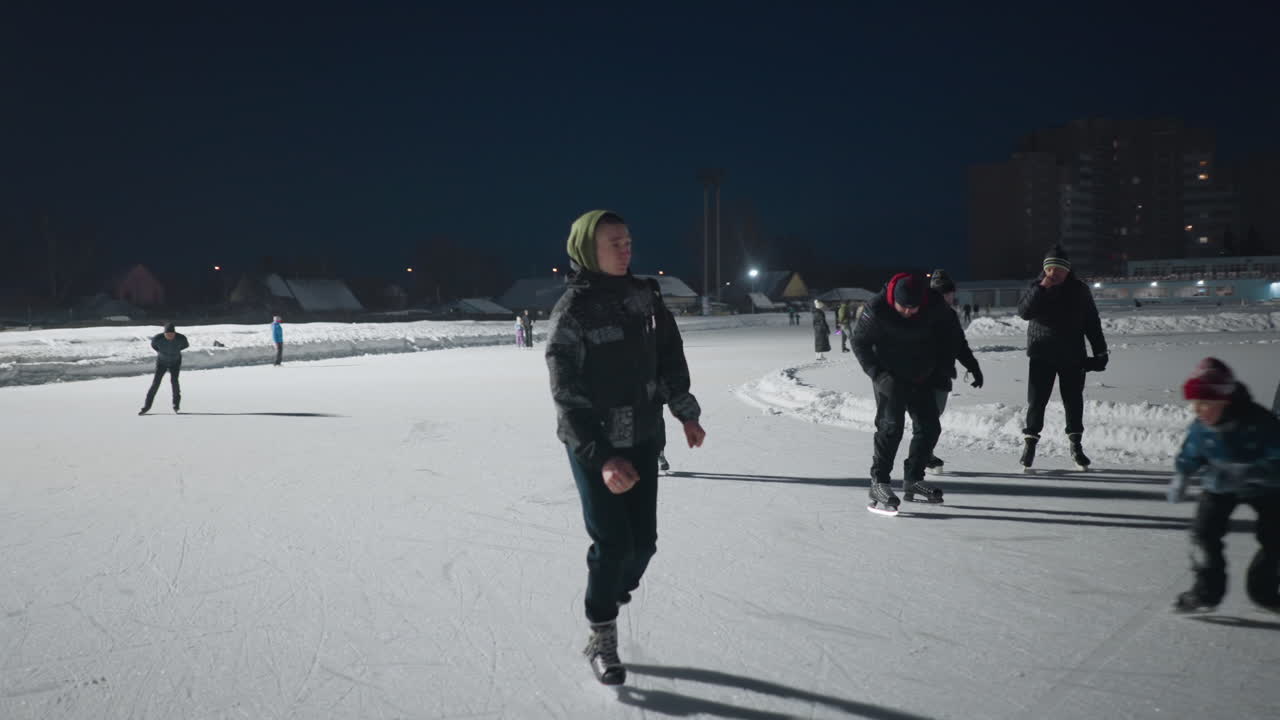 young people skating on spacious outdoor ice rink surrounded by snowbanks and city buildings at night illuminated by bright lights with lively crowd in background