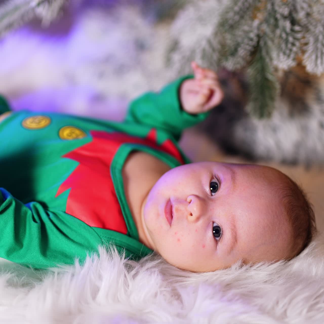 Active healthy baby in green elf costume lies on white fluffy plaid. Little kid waves his tiny arms and legs. Christmas tree at backdrop in blur