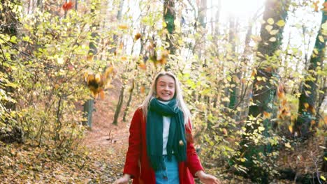 slowmotion young beautiful woman throwing leaves in the air amidst thee orange brown autumn forest woodland while wearing a red coat