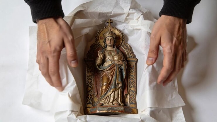 Entering frame, black-sleeved hands unwrapping tissue to reveal religious statue on white table