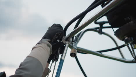 gloved hands attaching safety hook to metal frame of hot air balloon burner assembly during preflight setup under open sky with basket pumps hoses and regulators