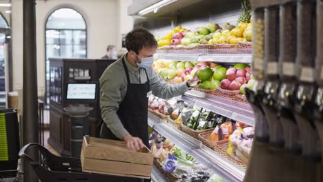 Busy-salesman-in-a-mask-is-putting-apples-on-shelves-at-fruits-section-in-supermarket-,-bearded-guy-is-wearing-apron