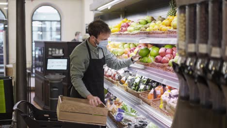 un vendedor ocupado con una máscara está poniendo manzanas en los estantes en la sección de frutas en el supermercado , un tipo barbudo está usando un delantal
