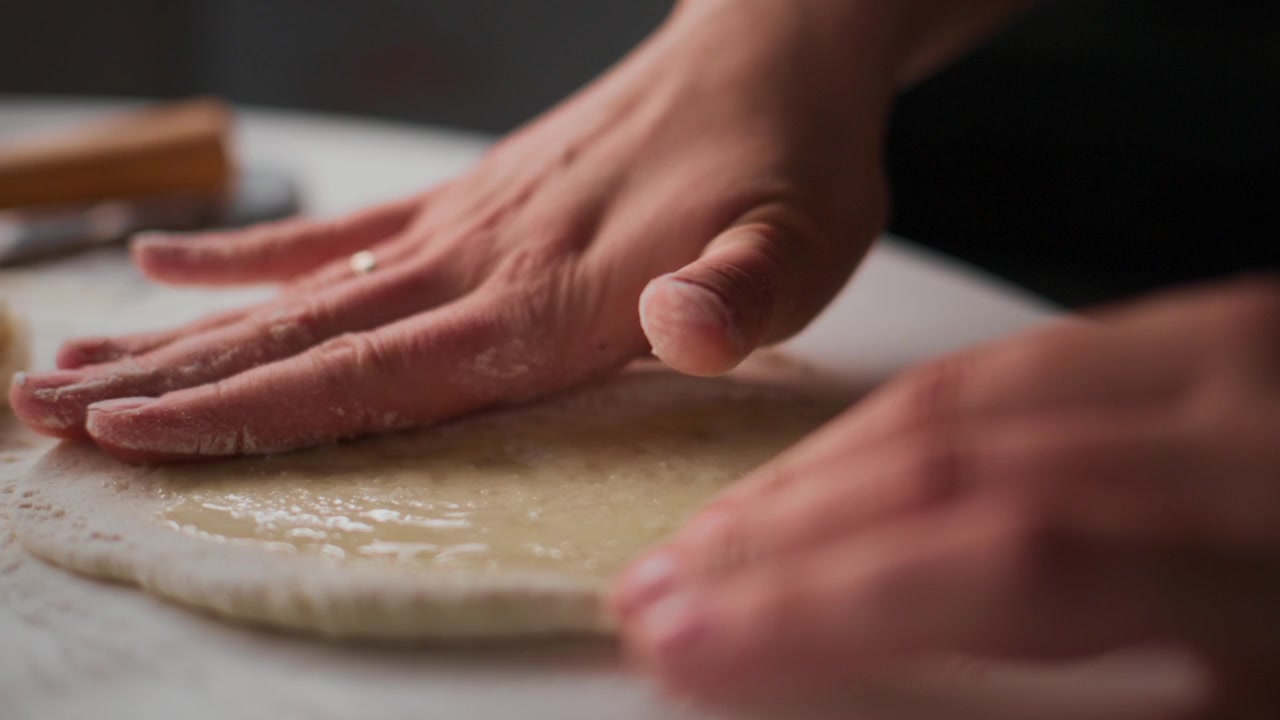 Primo piano di mani che premono delicatamente l'impasto su una superficie infarinata. Le dita stendono l'impasto in modo uniforme. La messa a fuoco morbida evidenzia la consistenza e il movimento. Ideale per contenuti di cucina, panificazione o preparazione alimentare.