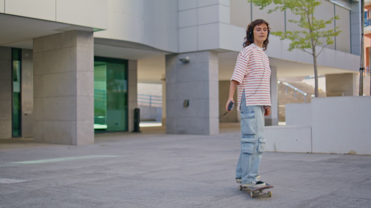 Relaxed girl riding skateboard at street. Woman skateboarding listening music