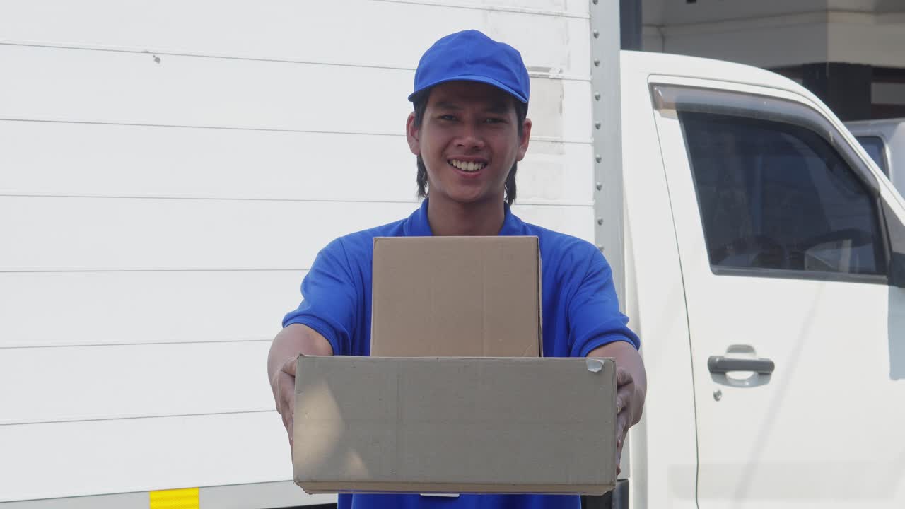 hombre de entrega asiático sonriente con gorra azul y camiseta sosteniendo una pila de caja de cartón, mostrándola y dándola a la cámara