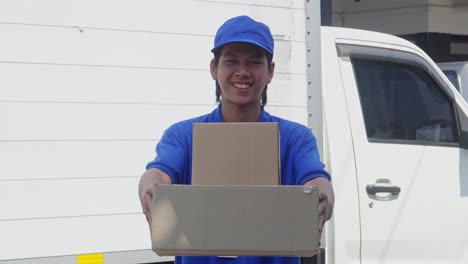 smiling asian delivery man wearing blue cap and t-shirt holding pile of cardboard box, showing and giving it to camera