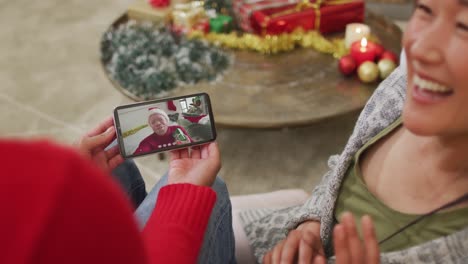 pareja asiática sonriente usando un teléfono inteligente para una videollamada de navidad con un hombre con sombrero de papá noel en la pantalla