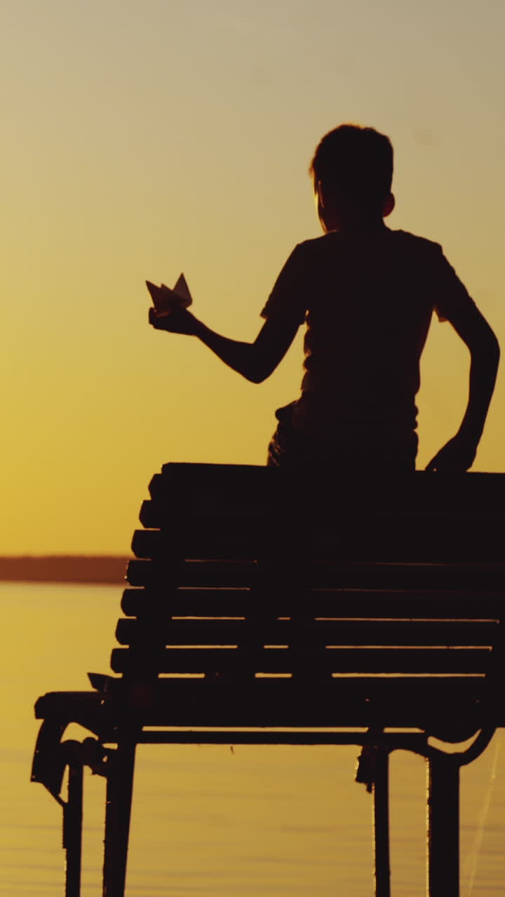 Little boy is playing with a paper boat on an old bridge by the river. Silhouette of little boy sitting alone at the river on sunset background Vertical video