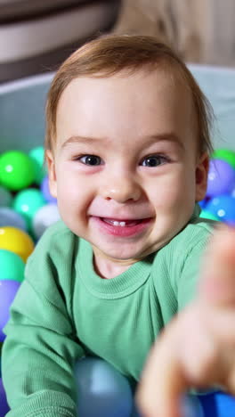 joyful happy toddler smiling broadly stands in soft basin filled with colorful balls. cheerful kid trying to reach the camera. close up. vertical video