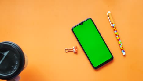 smart phone with green screen, coffee cup and pen on orange background