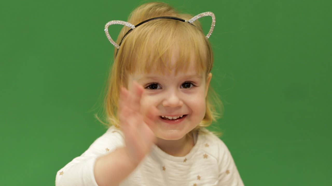 귀여운 아이는 미소 짓고 손을 흔들고 있습니다. 아름다운 2살 소녀. 귀여운 금발 아이. 갈색 눈. 가까운. 녹색 화면 배경