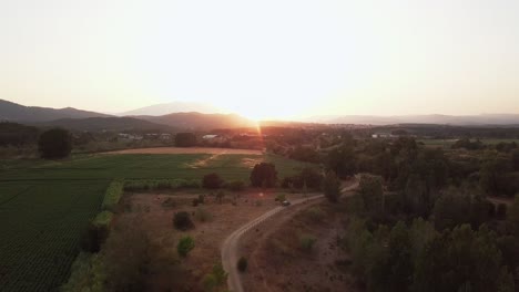 Aerial-drone-shot-flying-forward-over-a-gravel-road-with-green-fields-being-watered-and-an-amazing-sunset-in-the-back