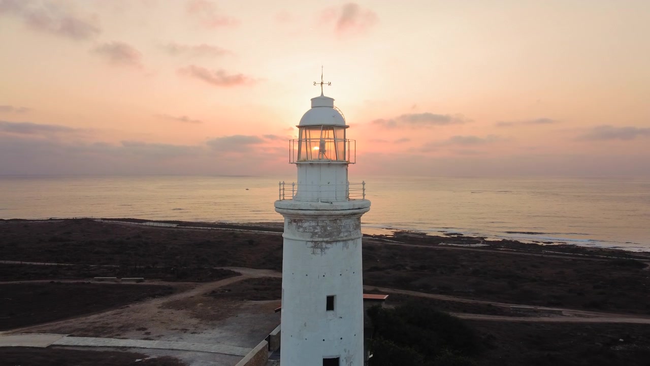 Alto faro bianco si erge a sentinella contro un cielo pastello mentre il sole tramonta sotto l'orizzonte, proiettando