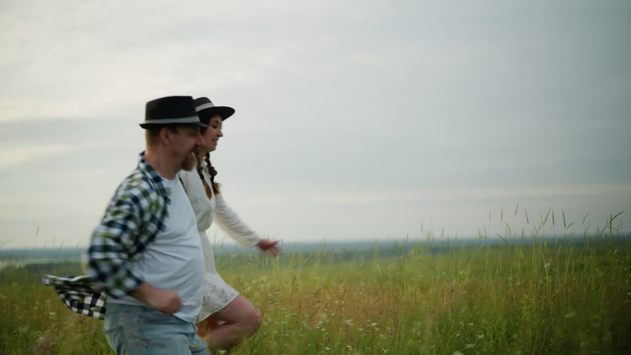 Partners hold hands and run through a grassy field at sunset, radiating joy and connection. The man wears a black hat and checkered shirt, while the woman is dressed in a white dress and matching hat