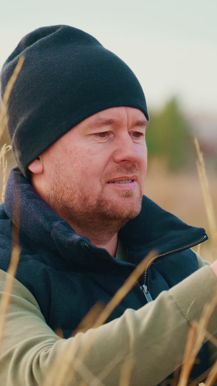 Un uomo vestito pesantemente con un berretto e un gilet è intensamente concentrato su ciò che lo circonda in un ambiente erboso all'aperto, trasmettendo contemplazione e connessione con la natura mentre esamina i dettagli del suo ambiente.