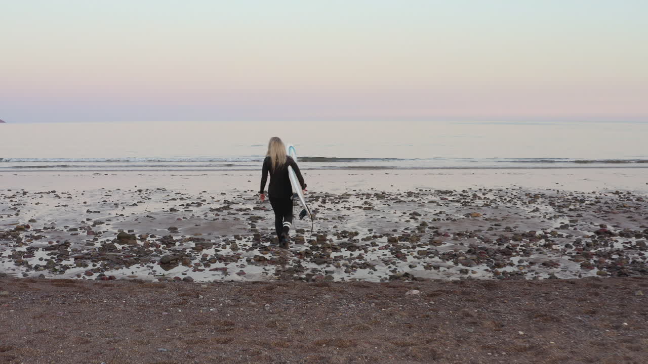 Donna di spalle in muta che trasporta una tavola da surf mentre entra in mare.