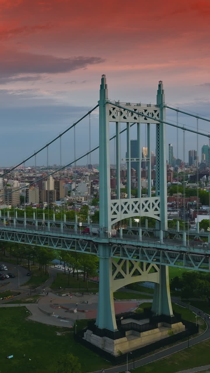 Triborough Bridge under the blue and red sky at sundown. Round parking lot and sports ground under the bridge. New York scenery at backdrop. Vertical video