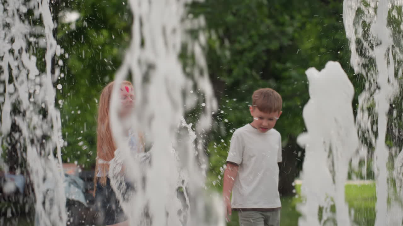 Cheerful kids playing with water fountain in recreational park, jets splashing around, summer greenery blurred behind, joyful movement and interaction capturing energetic outdoor fun