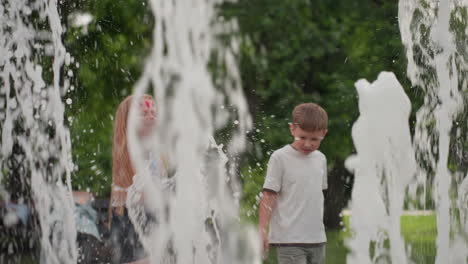 cheerful kids playing with water fountain in recreational park, jets splashing around, summer greenery blurred behind, joyful movement and interaction capturing energetic outdoor fun