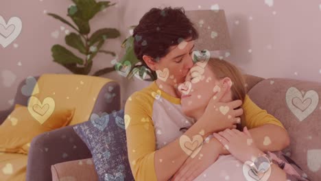animation of hearts over caucasian female couple sitting on sofa and embracing