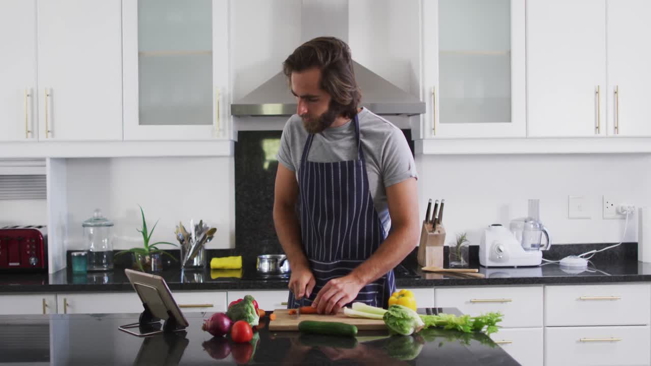 hombre caucásico con delantal cortando verduras mientras mira una tableta digital en la cocina