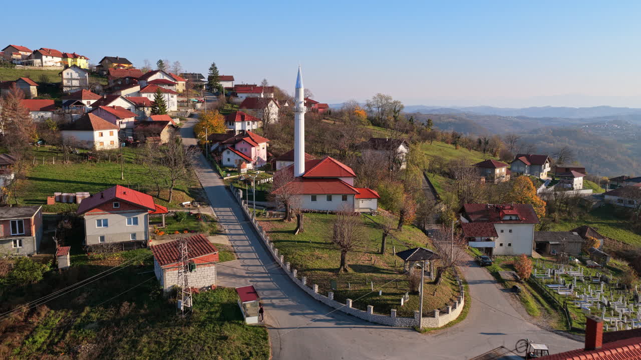 Aerial drone view of a picturesque hillside village featuring red roofed houses and a tall white mosque minaret, surrounded by open fields and distant mountain ranges in Bosnia and Herzegovina