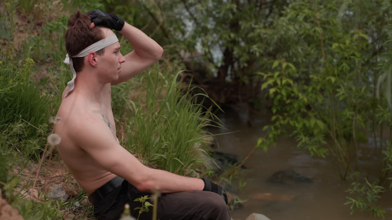 Side view of tourist with bare torso descending grassy slope near riverbank, pausing to sit while brushing hair with hand, wearing glove and headband, surrounded by lush greenery and natural daylight