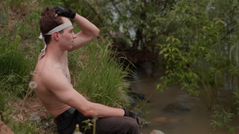side view of tourist with bare torso descending grassy slope near riverbank, pausing to sit while brushing hair with hand, wearing glove and headband, surrounded by lush greenery and natural daylight