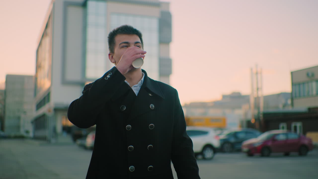 검은 코트를 입은 부동산 거장, 어두워지면서 밖에서 커피를 마시고, 행복한 표정, 건물, 주차된 자동차, 구조물을 특징으로하는 배경