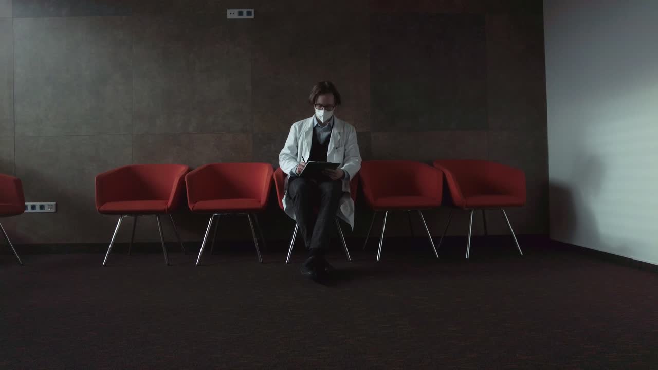 Psychologist with glasses in a white medical coat with a respirator sitting on a red chair making notes in a dark room