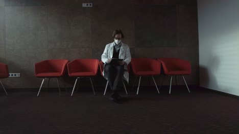 psychologist with glasses in a white medical coat with a respirator sitting on a red chair making notes in a dark room