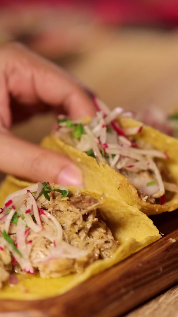 Hænder samler tacos med farverigt fyld på et træbræt under varm belysning på en restaurant