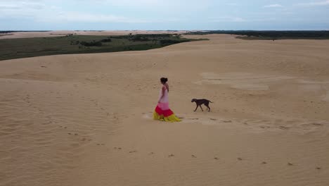 Una-Mujer-Con-Un-Vestido-Largo-Y-Fluido-Camina-Sobre-Una-Enorme-Duna-De-Arena-Con-Su-Perro