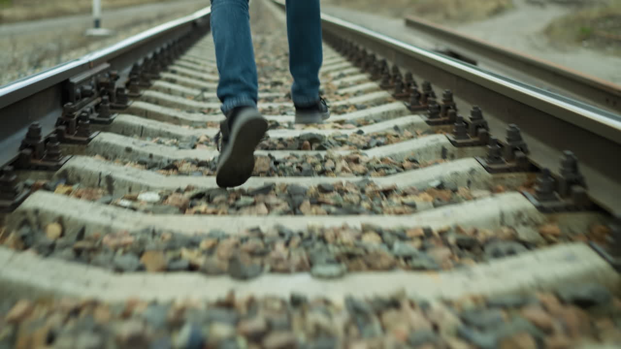A close view of someone running on a railway track, wearing jeans and canvas shoes, the track is covered with stones with a blur surrounding