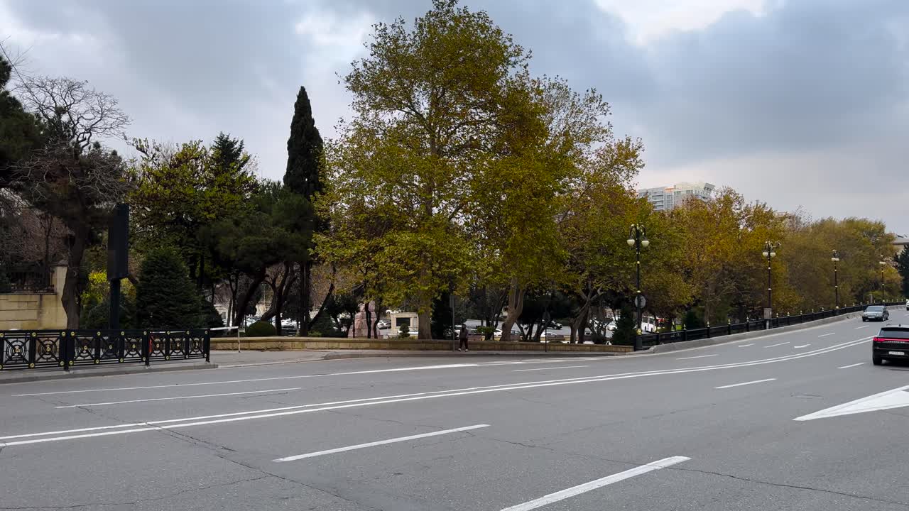 Cars drive past leafy autumn trees on a wide boulevard under cloudy skies. Baku, Azerbaijan