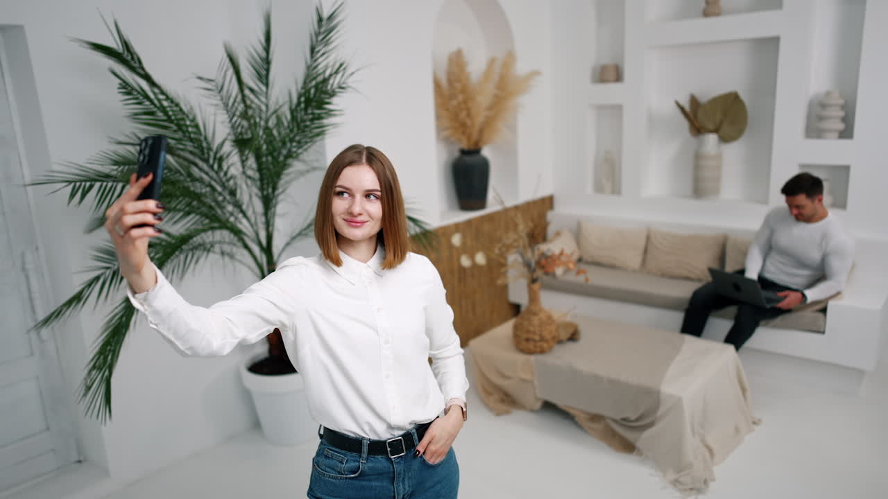 Positive Caucasian woman is taking selfie in the light cozy room. Lady points at man sitting on the sofa with laptop at backdrop.