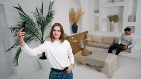 positive caucasian woman is taking selfie in the light cozy room. lady points at man sitting on the sofa with laptop at backdrop.