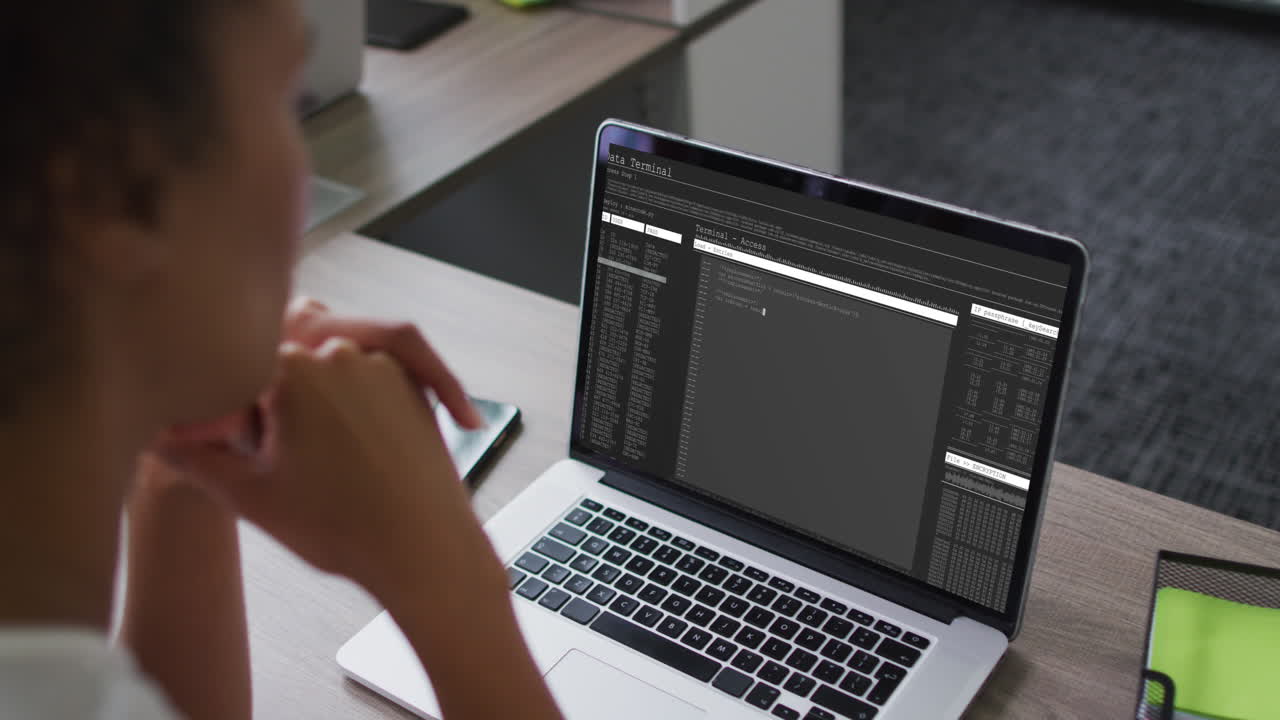 mujer biracial sentada en el escritorio viendo el procesamiento de datos de codificación en la pantalla de la computadora portátil